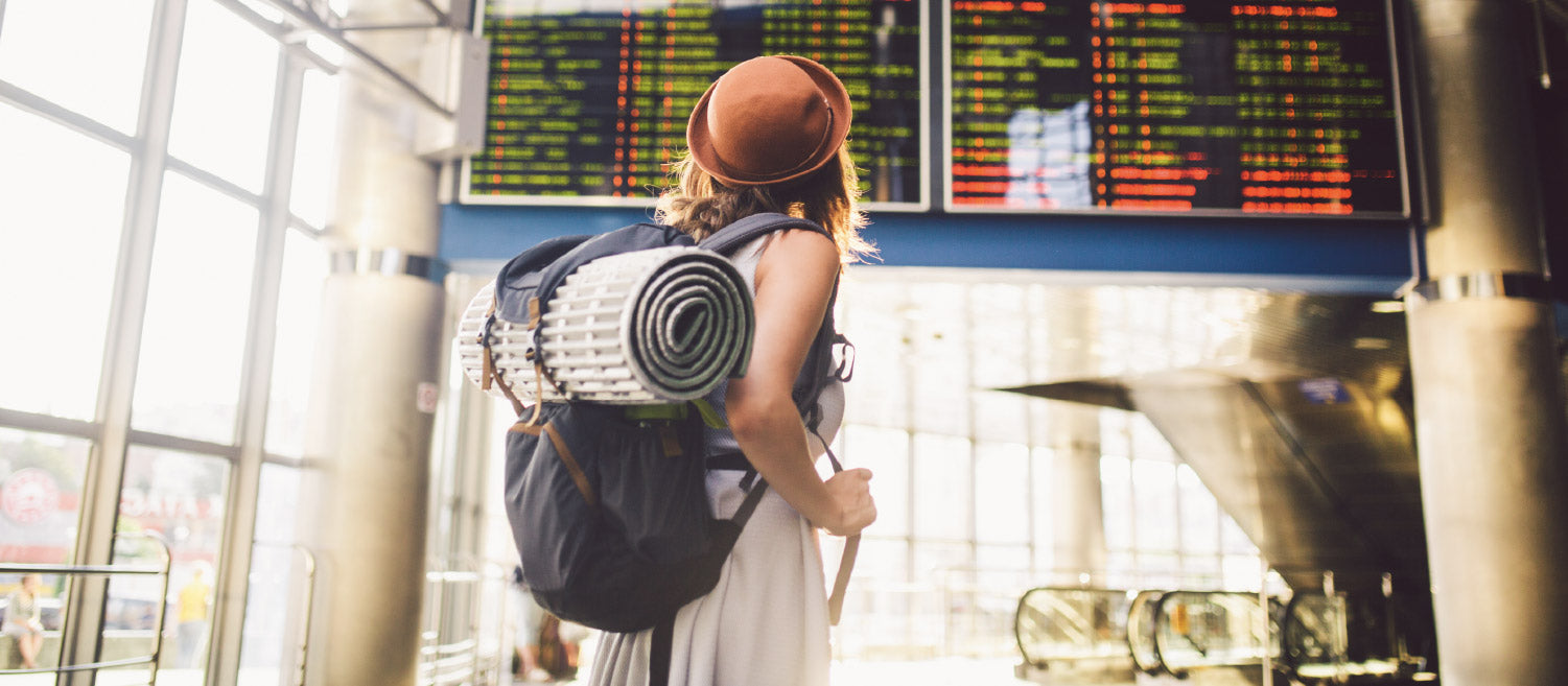 An adventure-seeking young women wearing a dress, hat and backpack looking up at a sign showing flight information at an airport