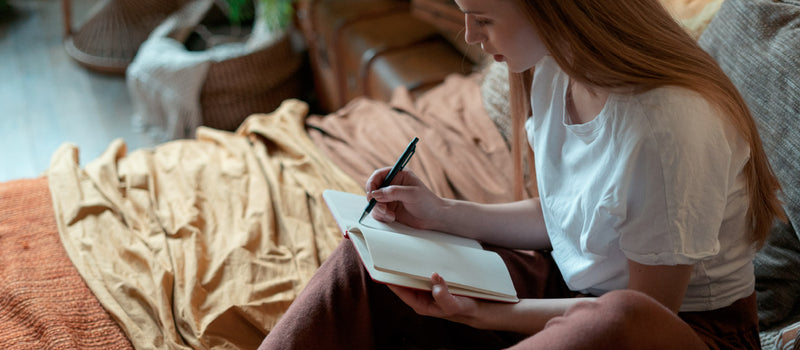 Woman with red hair sitting on her bed writing down her sleep affirmations in a journal to prepare for a restful sleep