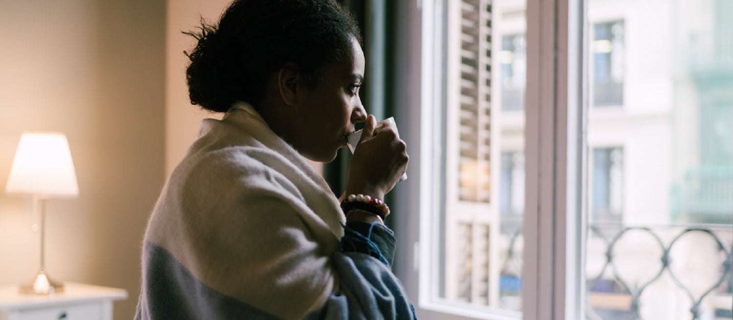 Women getting ready for bed by drinking tea, looking out the window, and wrapping a blue and white blanket around her shoulders