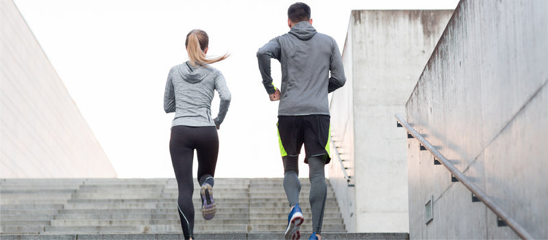 The backs of a man and women wearing workout clothing running together up a grey cement stairway with cement walls