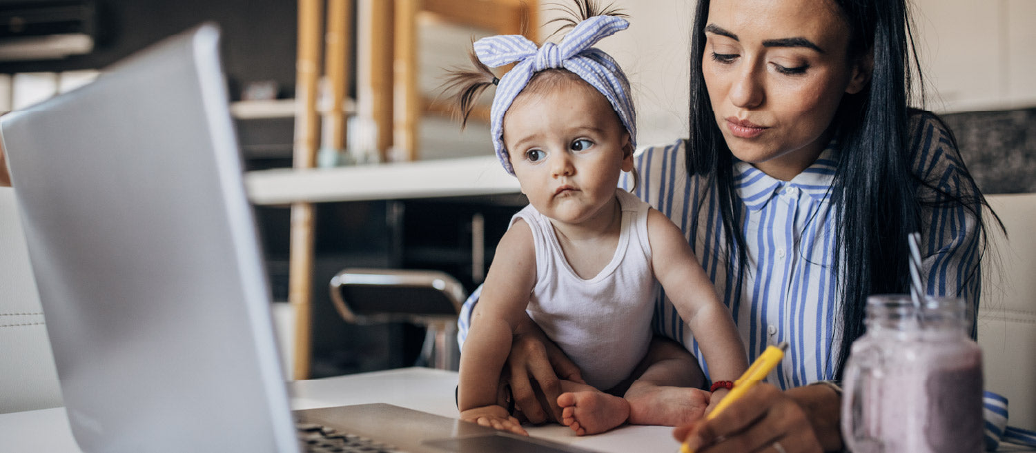 Closeup of a young mom wearing a blue striped shirt working at her desk while holding her daughter who has a bow in her hair