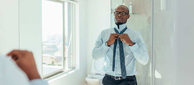 Man standing in his well-lit bathroom looking into a mirror and wearing glasses a blue button-down shirt dress pants and a tie