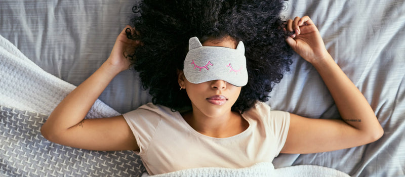 Sleeping women with her arms near her head wearing a gray eye mask with pink sewn on eyelashes showing the benefits of beauty sleep