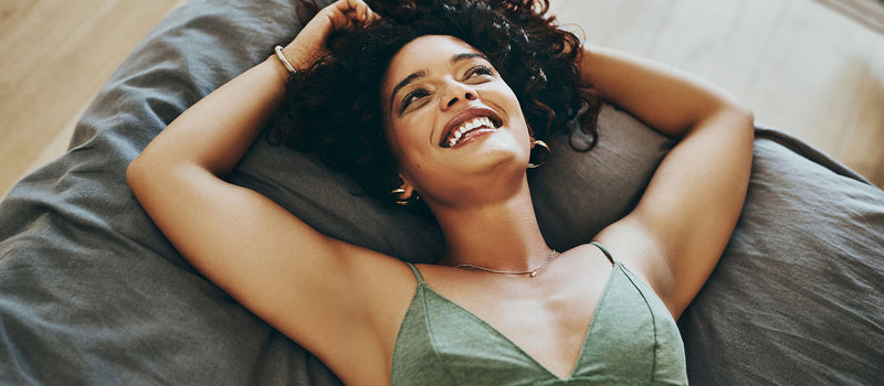 Women with dark hair preparing to sleep in a bra by laying on a bed with grey sheets looking up and smilng