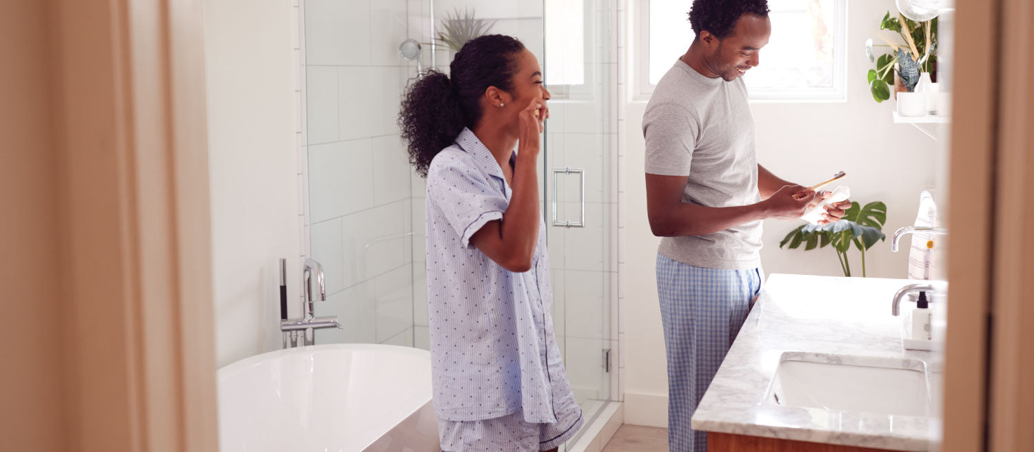 Man and woman brushing their teeth in their brightly lit bathroom while wearing different types of pajamas